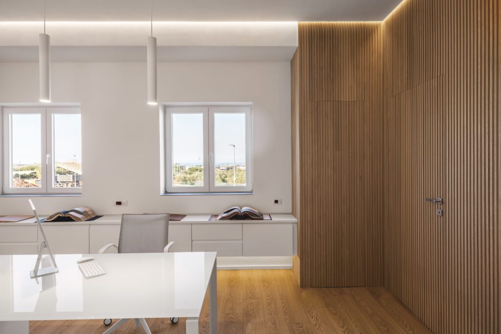 La porta di comunicazione tra l'ufficio direzionale e l'amministrazione è una filo muro rivestita con pannellatura a doghe in rovere naturale. Il profilo led nascosto illumina a cascata le doghe in legno per un effetto caldo e soft.