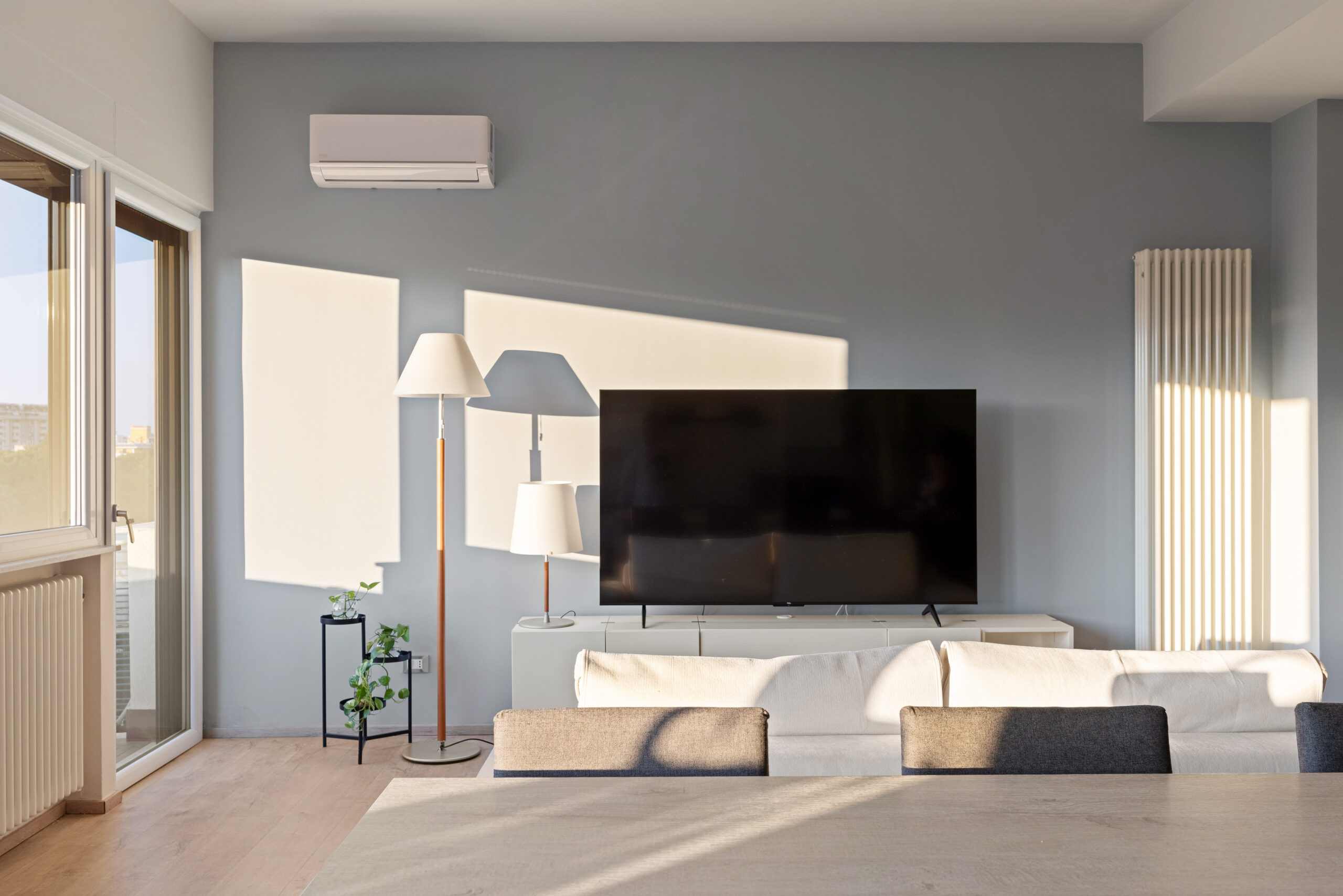 Dettaglio della parete TV grigia nel soggiorno con forti contrasti di luce naturale geometrica, lampada da terra Artemide con stelo in legno, climatizzatore e mobile basso lineare negli Appartamenti Parco Trabia.