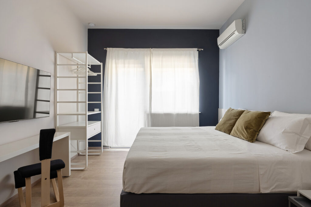 Interno della camera da letto matrimoniale con letto tessile color rosa antico, cuscini d'accento, armadio a giorno bianco e climatizzatore a parete negli Appartamenti Parco Trabia.