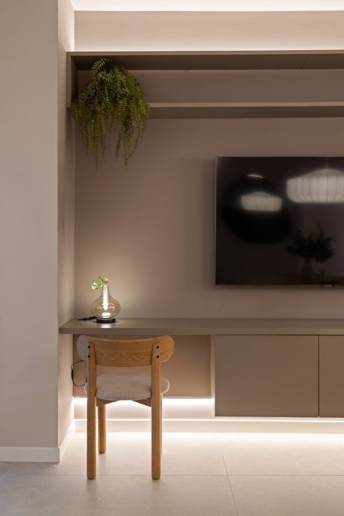 Primo piano della parete TV attrezzata con mensola sospesa, TV a schermo piatto, lampada da tavolo decorativa e sedia in legno nella struttura ricettiva di lusso a Palermo.